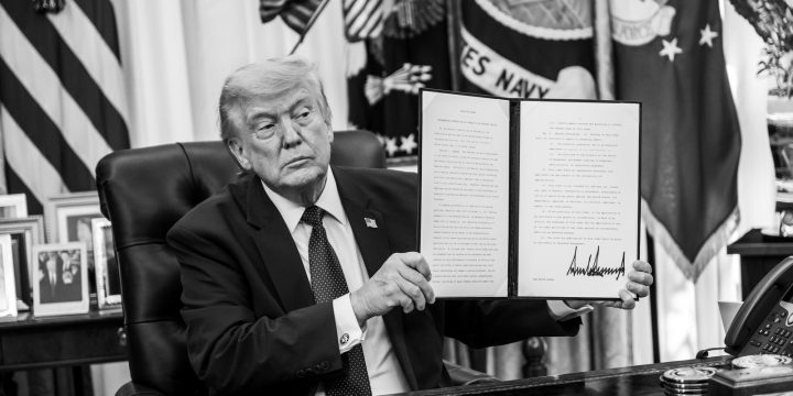 President Trump seated at the Oval Office desk holding a signed document