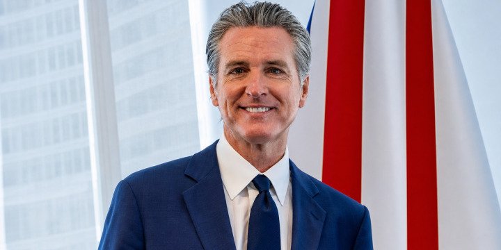 California Governor Gavin Newsom in a blue suit beside an American flag