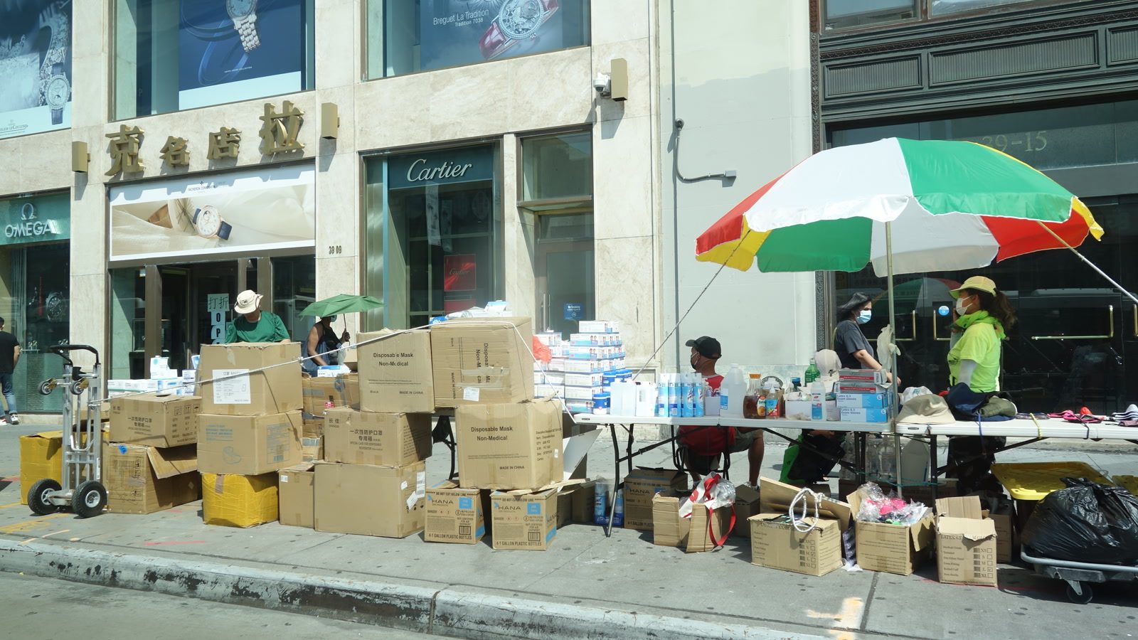 Sidewalk vendors display boxed goods and bottles on folding tables under an umbrella in New York City