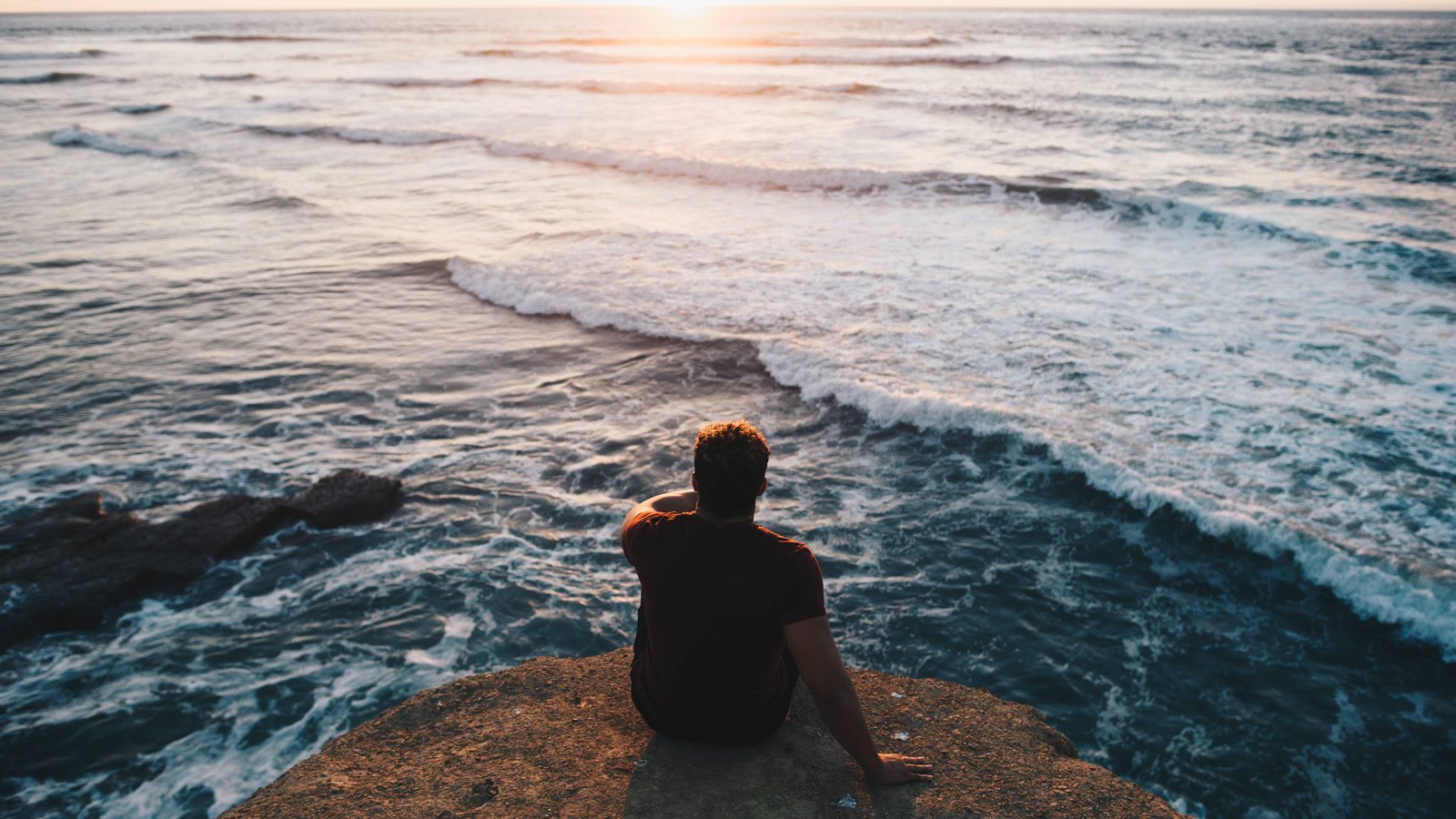 A visitor sits on a rocky ledge above Pacific waves at Sunset Cliffs in San Diego
